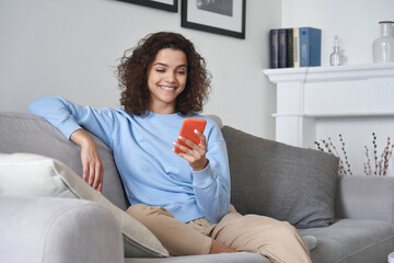 Happy 20s young woman watching social media videos looking at smartphone relaxing on couch, smiling...
