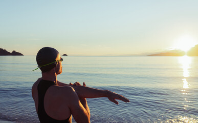 Back view of fit male swimmer in swimsuit and cap standing on seashore and stretching arms before training
