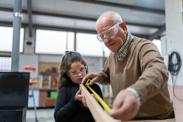 Adorable little girl in casual clothes and goggles helping grandfather measuring wooden board with tape during work in manufacture