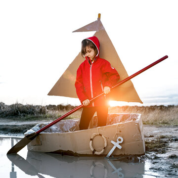 Cute kid with paddle playing in handmade cardboard boat while entertaining in countryside at weekend