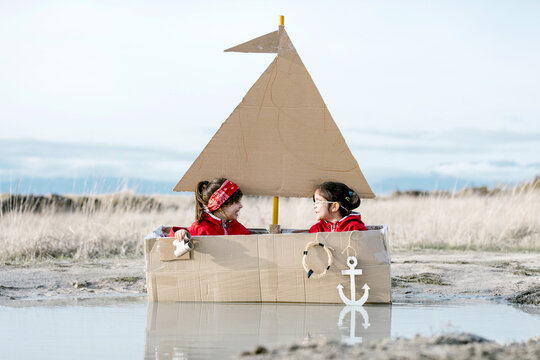 Playful Children Sitting In Handmade Cardboard Boat In Puddle And Looking Through Spyglass While Having Fun At Weekend Together