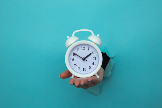 Woman hand holds alarm clock through a blue paper hole. Time management and deadline concept.