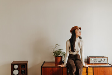 Attractive girl in velvet pants and caps sits on chest of drawers against white wall