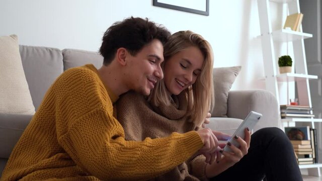 Young happy couple having fun holding smartphone using mobile phone at home, laughing, playing game, doing online shopping in ecommerce app together, checking social media applications together.