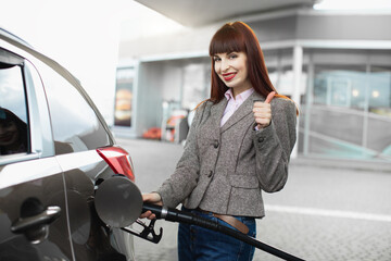 Attractive cheerful young business woman, smiling and showing her thumb up to camera, while refueling her luxury car at gas station