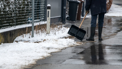 Mann in Gummistiefeln beim Schneeschaufeln auf einem Gehweg