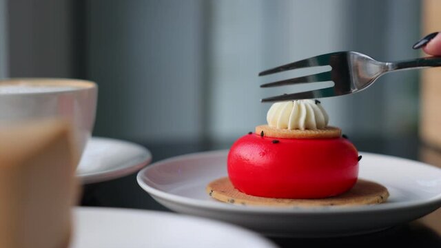 A hand cuts a piece of cake with a fork. A piece of red glazed mousse cake on a fork. Sponge cake and spongy dessert. Desserts are red. Composition of a sweet snack in a cafe. Aromatic cappuccino
