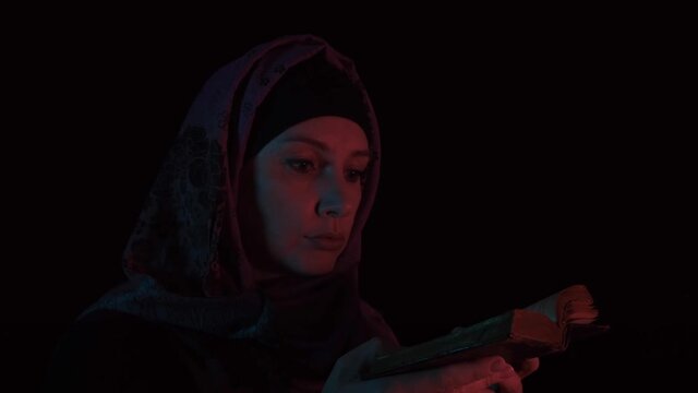 A woman in headscarf opening an old Holy Bible in the dark at night and praying to God