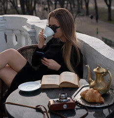 Sitting on a balcony above a European street, a girl with long blond hair is drinking tea or coffee. Business meeting, breakfast or lunch