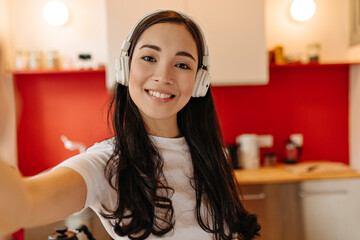 Brown-eyed woman in white headphones smiles and takes selfie in kitchen