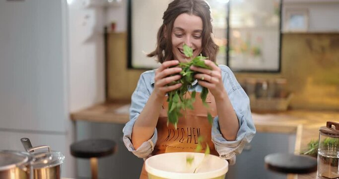 Young and happy housewife mixing green salad in the bowl on the kitchen table. Healthy vegan food concept