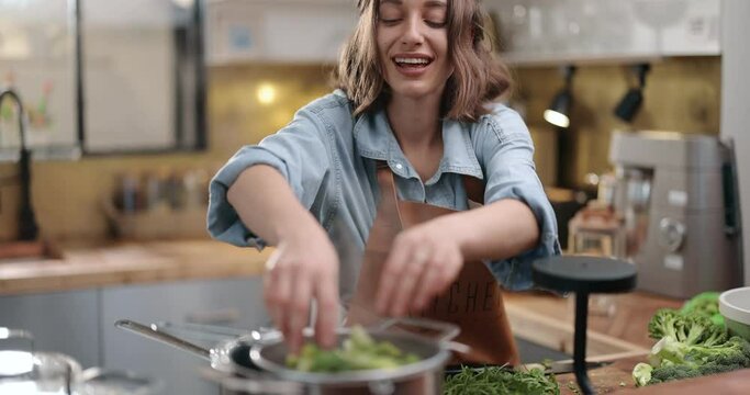 Young cheerful housewife in apron cooking broccoli on the kitchen at home. Happy household chores and healthy eating concept