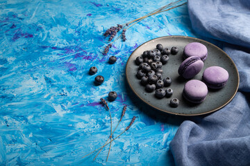 Purple macarons or macaroons cakes with blueberries on ceramic plate on a blue concrete background. Side view, copy space.