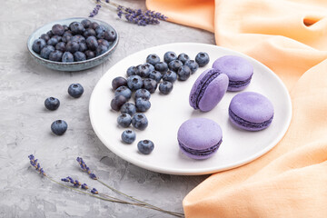 Purple macarons or macaroons cakes with blueberries on white ceramic plate on a gray concrete background. Side view.
