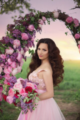 Fototapeta premium Beautiful bride in pink dress standing with her bouquet at a round flower arch under a large single tree in a field at sunset. A destination summer wedding, romantic country ceremony.