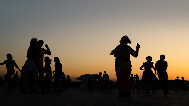Many unrecognizable people silhouettes dancing on city embankment against sky at sunset - slow motion. Warm illumination, evening time. Romantic pairs group dance concept