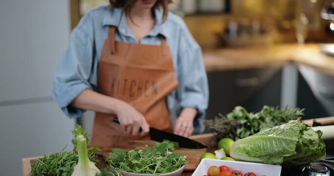 Young cheerful housewife making salad on a kitchen table full of healthy green food ingredients. Vegan organic food, healthy cooking concept