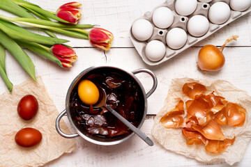 the process of painting Easter eggs with natural vegetable dyes, onion husks, on a white wooden background top view of chicken eggs and red tulips