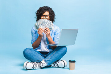 Happy young curly african american beautiful woman sitting on the floor with crossed legs and using laptop on blue background. Holding money bills.