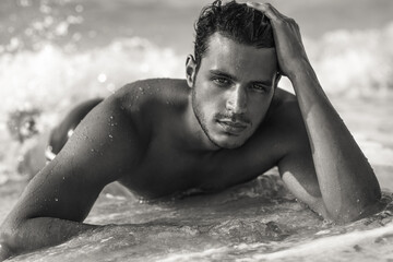 Portrait of sexy handsome topless male model with beautiful eyes laying in the ocean water on the beach. Black and white.