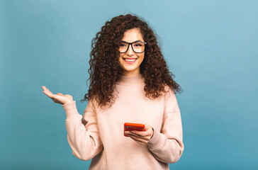 Portrait of a cheerful casual curly girl holding a mobile phone and pointing her finger away from isolated on blue background.