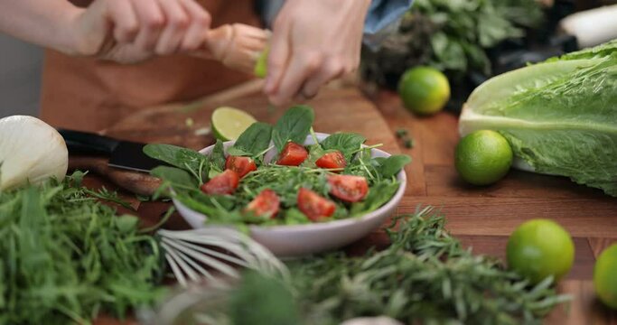 Housewife squeezing lemon juice on the salad, preparing healthy food on the kitchen table with green veggie ingredients. Close-up slow motion shot