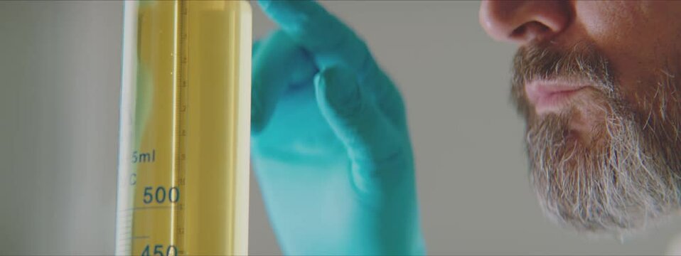 Close-up of graduated cylinder full of yellow liquid. Man reading measured value from scale. Working in factory laboratory concept.