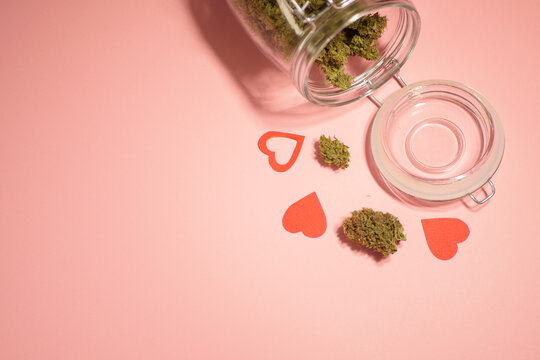 Cannabis buds in glass jar. Pink background. Valentine's day. Paper hearts. Copy space