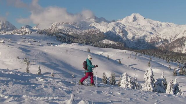AERIAL: Young female tourist hikes down a snowy hill during a ski touring trip to scenic Velika Planina, Slovenia. Splitboarder explores the picturesque Slovenian mountains on a sunny winter day.