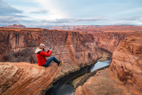 Man taking picture with mobile phone while sitting on a cliff over Colorado river in Horseshoe bend canyon