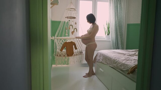 Young pregnant girl hangs baby clothes on a crib with toys