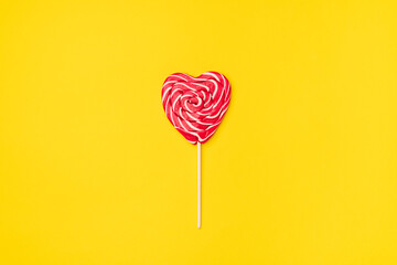 Red heart-shaped lollipop candy isolated on an empty colorful yellow background. Symbol of love for Happy Women's, Valentine's Day, Birthday greeting romantic postcard. Flat lay, top view, copy space