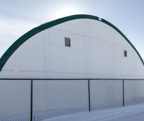 Large fabric quonset hut hanger at an airport chain link fence in foreground snow and sun flare...