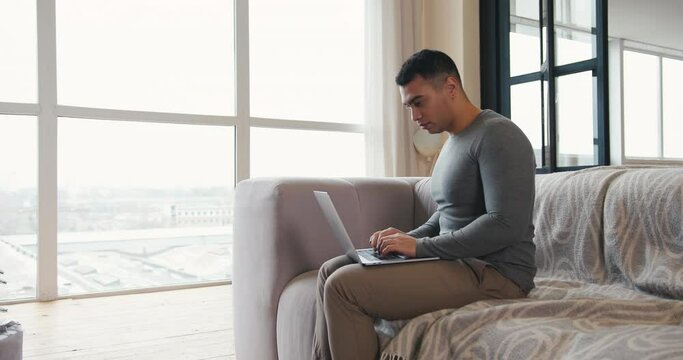 Young brunette man in grey shirt surfs internet with contemporary laptop sitting on comfortable sofa in living room slow motion