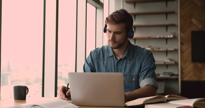 Focused millennial male student wearing headphones, listening educational lecture, preparing for exam online using electronic and paper books, writing notes, improving knowledge, e-learning concept.