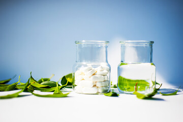 Two glass jars with pills on a blue background and a white table. Green leaves and white pills. Health theme.