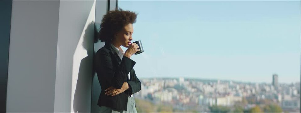 Businesswoman drinking coffee at the office and looking out the window of high rise skyscraper building during morning tea break. 