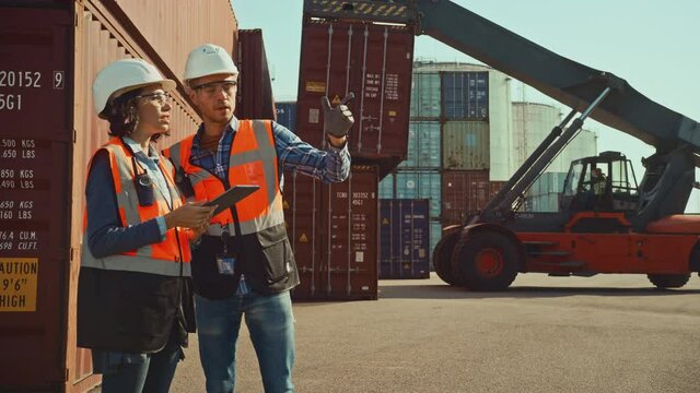 Multiethnic Female Industrial Engineer with Tablet Computer and Male Foreman Worker in Hard Hats and Orange High-Visibility Vests Stand in Container Terminal. Colleagues Talk About Logistics Business.
