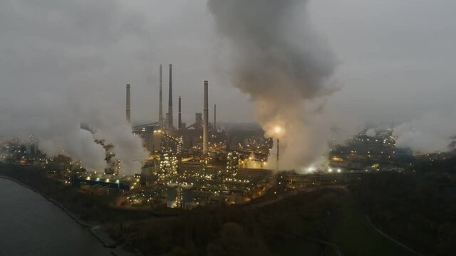 Aerial view of illuminated petrochemical factory during nightfall. White smoke polluting the air
