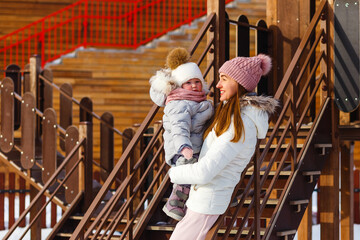 a young happy mother and a little girl in warm outerwear walk in the park in the playground in...