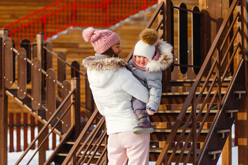 a young happy mother and a little girl in warm outerwear walk in the park in the playground in winter. beautiful woman and baby in jackets