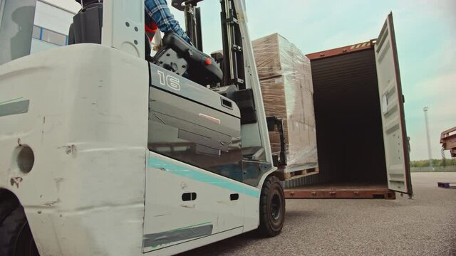 Forklift Driver Loading a Shipping Cargo Container with a Full Pallet with Carboard Boxes in Logistics Operations Port Terminal. Tracking Shot from the Side Shows How Pallet is Brought by the Machine.