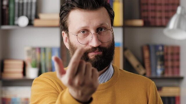 Man beckons with his finger. Serious frowning bearded man with glasses in office or apartment room looking at camera and points his fingers, gesturing to him. Close-up and slow motion