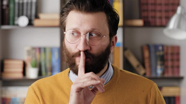 Shh, man secret finger. Suspicious bearded man in glasses in office or apartment room looking at camera and brings his index finger to his mouth lips and she say shhh. Close-up and slow motion