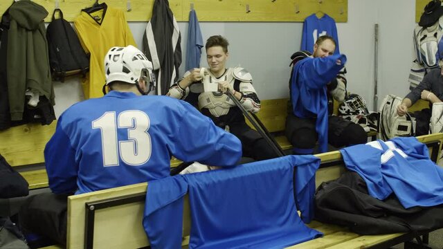 Professional male hockey players sitting in locker room, putting on uniform, preparing sticks and chatting before game
