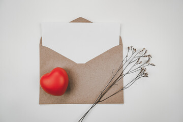 Blank white paper is placed on open brown paper envelope with red heart with Limonium dry flower on white background. Valentine's day concept.