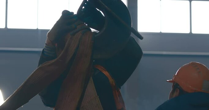 A worker inside a hangar under construction puts textile slings on a crane hook to lift a heavy load 