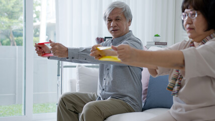 Young older senior asia citizen couple sit on sofa couch with in-home nursing care, assisted living, scrubs nurse use resistance band exercise for senior patient in physiotherapy treatment at home.