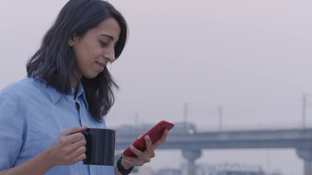 a young indian woman entrepreneur standing outdoors holding a coffee mug in one hand and using mobile phone with other hand , with a blurry background of city metro train passing by