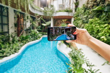 Hand of woman photographing swimming pool in spa resort on smartphone to upload photo on social...